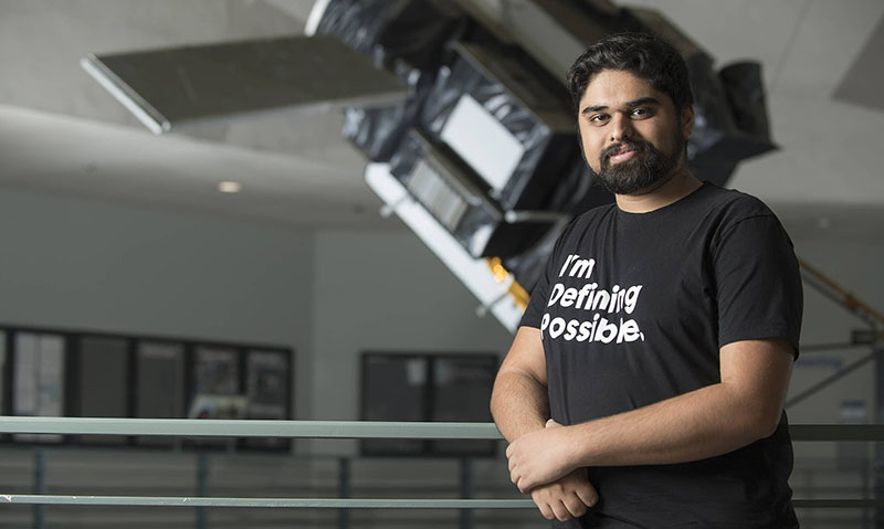 A man with a black t-shirt that reads "I'm Defining Possible" and a model satellite behind him.